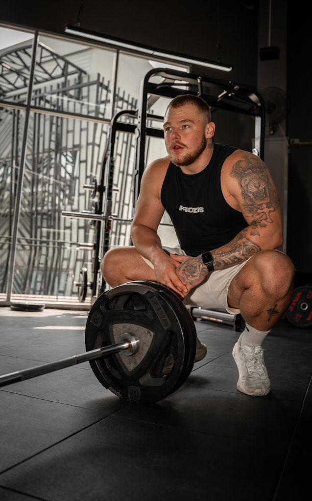 Trainer sitzt auf dem Boden neben einer Langhantel im Fitnessstudio und blickt zur Seite.